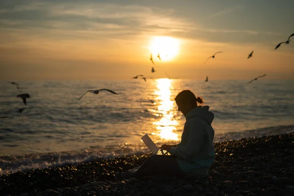 Femme blanche tapant sur un ordinateur portable au bord de la mer au coucher du soleil. Travail indépendant. — Photo