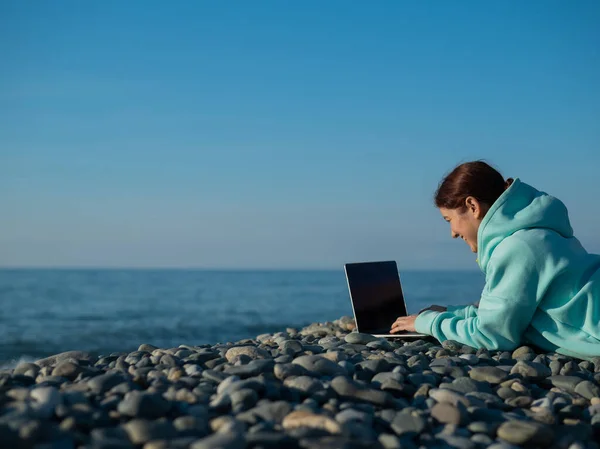 Mulher caucasiana deitada de barriga em uma praia de seixos e digitando em um laptop. Freelancer de trabalho remoto. — Fotografia de Stock