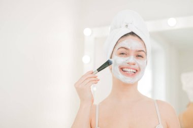 Saçında havlu olan güzel beyaz kadın kilden bir maske takıyor. Evdeki güzellikle ilgilenmek..