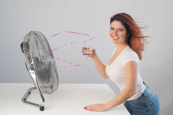 Una mujer caucásica pelirroja se enfría junto al ventilador eléctrico y bebe una bebida fría. Climatización en el apartamento — Foto de Stock