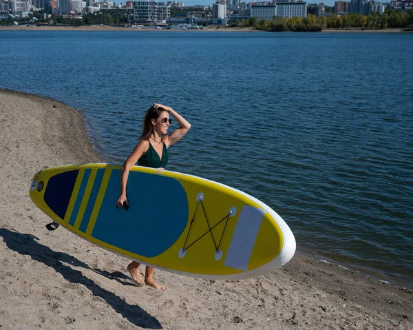 Mujer caucásica camina a lo largo de la playa y lleva un sup board en el río en la ciudad. Deportes de verano. — Foto de Stock