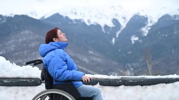 Žena na invalidním vozíku je chladná a dýchá do dlaní, aby se zahřála ve zasněžených horách.. — Stock video