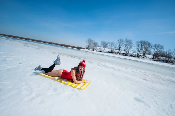 Кавказька жінка в червоному купальнику і в'язані капелюхи сонячні ванни взимку лежать на снігу. — стокове фото
