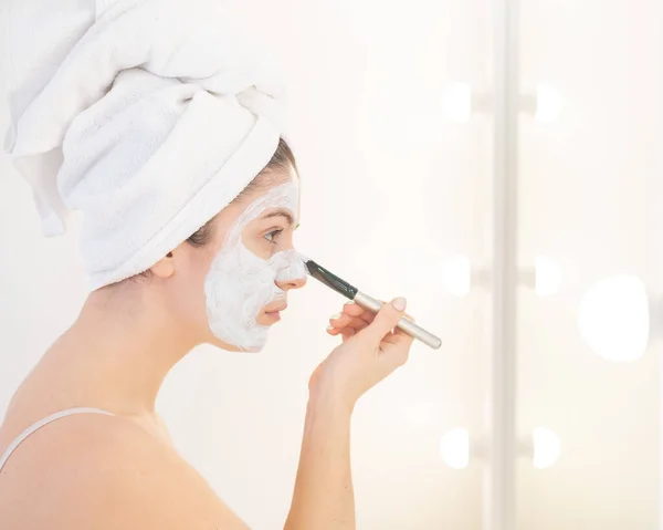 Hermosa mujer caucásica con una toalla en el pelo se aplica una mascarilla de barro. Cuidar de la belleza en casa — Foto de Stock