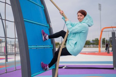 Nane rengi kazaklı beyaz bir kadın açık hava spor sahasında dar bir ipe tırmanıyor.. 