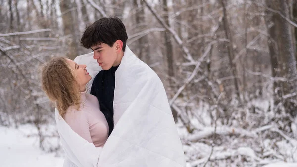 Um jovem casal caminha no parque no inverno. O cara e a menina estão se beijando envolto em um cobertor branco ao ar livre. — Fotografia de Stock