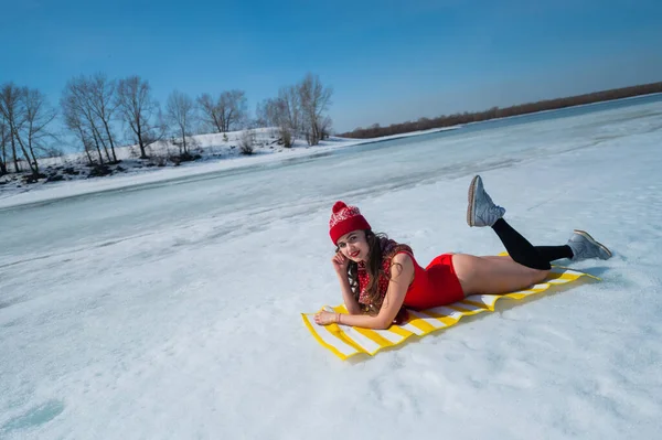 Mulher branca em um maiô vermelho e um chapéu de malha banhos de sol no inverno deitado na neve. — Fotografia de Stock