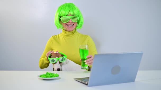 Młoda kobieta w zielonej peruce i wesołych okularach pije piwo i gryzie oszklone orzechy. Dziewczyna siedzi z psem przy stole i świętuje dzień Św. Patryka online czatując z przyjaciółmi na laptopie. — Wideo stockowe