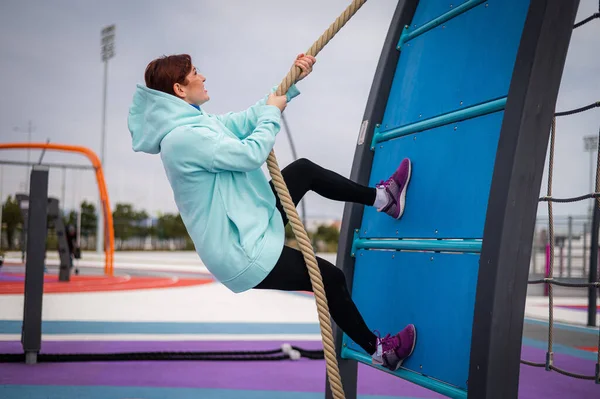 Λευκή γυναίκα με φούτερ από μέντα σκαρφαλώνει σε τεντωμένο σχοινί σε υπαίθριο χώρο άθλησης.. — Φωτογραφία Αρχείου