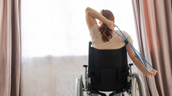 Blanke vrouw in een rolstoel doet oefeningen met behulp van fitness elastiekjes. — Stockfoto