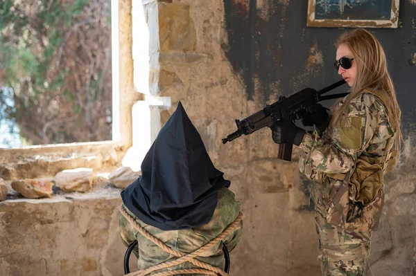 Una donna caucasica in uniforme militare tiene un ostaggio con una borsa in testa alla vista di una mitragliatrice. — Foto Stock