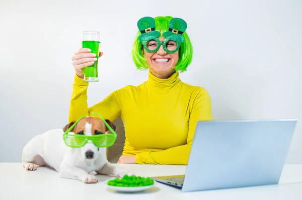 En ung kvinna i grön peruk och glada glasögon dricker öl och biter glaserade nötter. En flicka sitter med en hund vid ett bord och firar st patricks dag online chatta med vänner på en bärbar dator. — Stockfoto