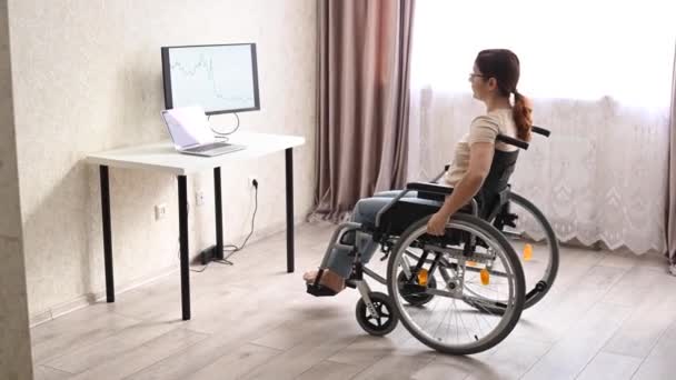 En hvid kvinde i en kørestol kører op til sit skrivebord og begynder at skrive med en bærbar computer hjemmefra. Fjernarbejde for handicappede. – Stock-video