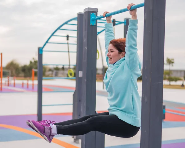 Kaukasische Frau im Mint-Sweatshirt hängt an einer waagerechten Stange und trainiert auf dem Sportplatz im Freien die Bauchmuskeln. — Stockfoto