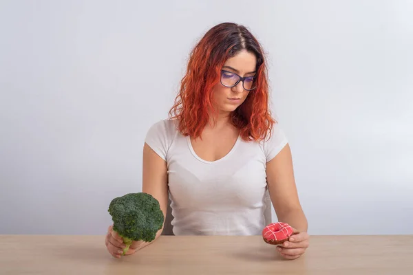 Kaukasisk kvinna som drömmer om snabbmat. rödhårig flicka väljer mellan broccoli och munkar på vit bakgrund. — Stockfoto