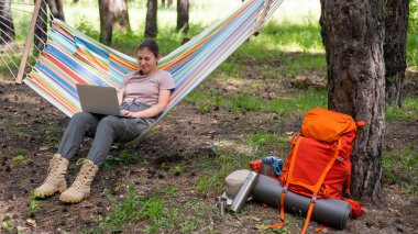 Ormandaki bir hamakta otururken dizüstü bilgisayarda çalışan beyaz bir kadın. Yürüyüşte kablosuz bilgisayar kullanan bir kız..