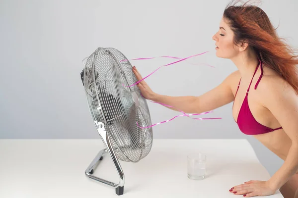 Red-haired smiling woman in a bikini drinks a cold drink and enjoys the blowing wind from an electric fan on a white background. Climate control on a hot summer day