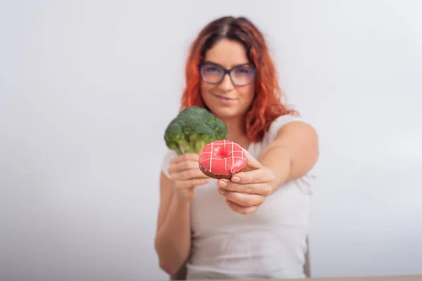 Běloška si vybírá mezi zeleninou a rychlým občerstvením. Zrzka dívka drží brokolici a koblihu na bílém pozadí. — Stock fotografie