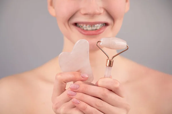 Retrato de cerca de una mujer con frenos en los dientes sosteniendo un masajeador rodillo rosa y un raspador gouache sobre un fondo blanco. —  Fotos de Stock