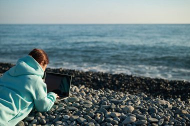 Beyaz bir kadın, çakıl taşı plajında yüzüstü yatıyor ve dizüstü bilgisayarında yazı yazıyor. Uzak çalışma serbest çalışanı.