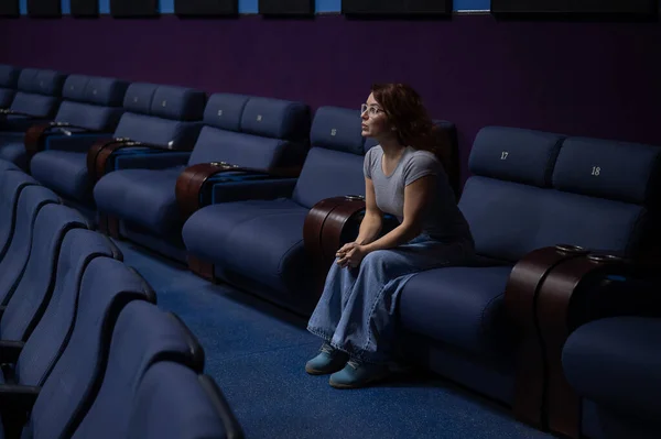 Caucasian red-haired woman sits on the last row in a cinema in an empty hall. The girl is watching a movie alone.