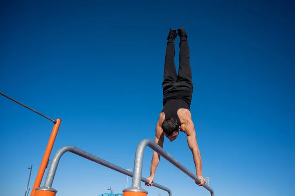 Hombre sin camisa haciendo handstand en barras paralelas en el campo de deportes. —  Fotos de Stock