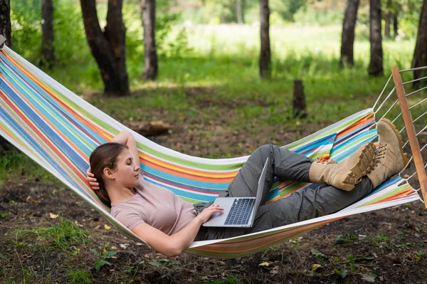 Mulher caucasiana trabalhando em laptop enquanto sentada em uma rede na floresta. Menina usa um computador sem fio em uma caminhada. — Fotografia de Stock