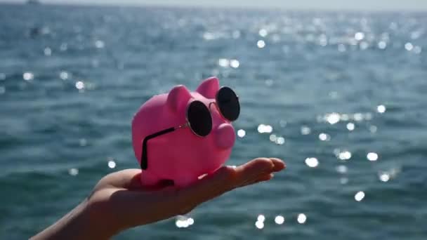 Eine Frau hält ein Sparschwein mit Sonnenbrille vor dem Hintergrund des Meeres. Haushaltsferien. — Stockvideo