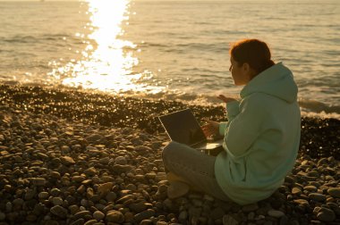 Beyaz kadın, çakıl taşlı plajda dizüstü bilgisayardan video ile iletişim kuruyor.. 