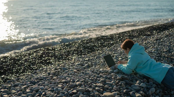 Кавказька жінка лежала на животі на гальному пляжі і друкувала на ноутбуці. Дистанційна робота фрілансер. — стокове фото