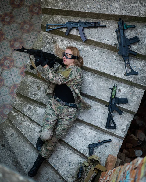 Caucasian woman in military uniform lies on the stairs of an abandoned building and holds a machine gun. View from above.