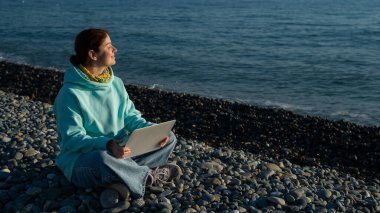 Bir çakıl taşı kumsalında otururken dizüstü bilgisayarda çalışan mutlu beyaz kadın..