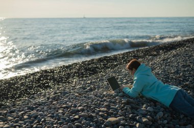 Beyaz bir kadın, çakıl taşı plajında yüzüstü yatıyor ve dizüstü bilgisayarında yazı yazıyor. Uzak çalışma serbest çalışanı.