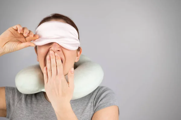 Caucasian woman yawns with travel pillow and sleep mask on white background.