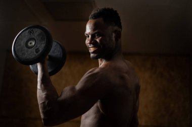 Çekici Afrikalı Amerikalı adam gülümsüyor ve halterlerle egzersiz yapıyor.. 
