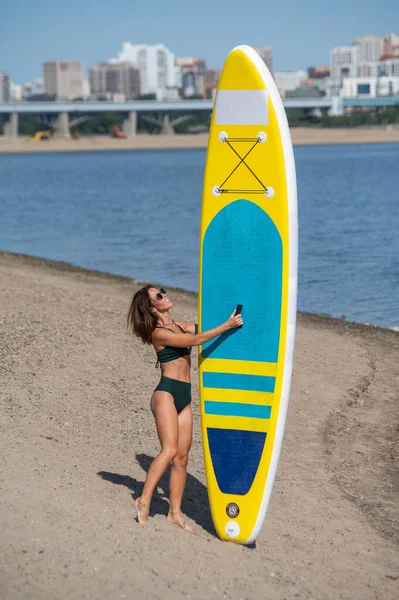 Mujer caucásica camina a lo largo de la playa y lleva un sup board en el río en la ciudad. Deportes de verano. —  Fotos de Stock