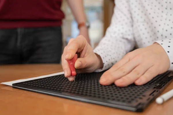 A man teaches a woman how to use a special stencil and stylus to write a letter in Braille.