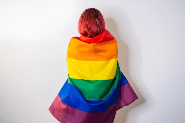 Young red-haired woman stands with her back against a white background and holds the lgbt flag — Stock Photo, Image