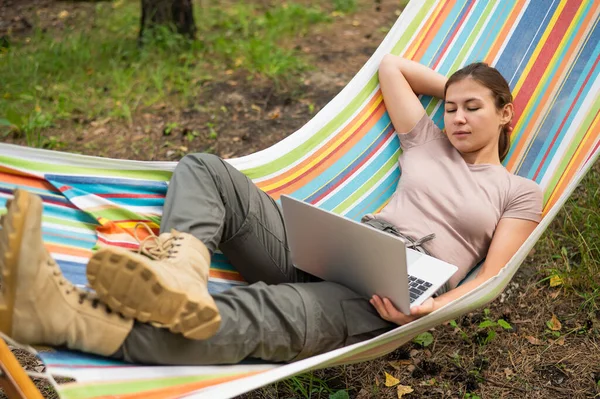 Mulher caucasiana trabalhando em laptop enquanto sentada em uma rede na floresta. Menina usa um computador sem fio em uma caminhada. — Fotografia de Stock