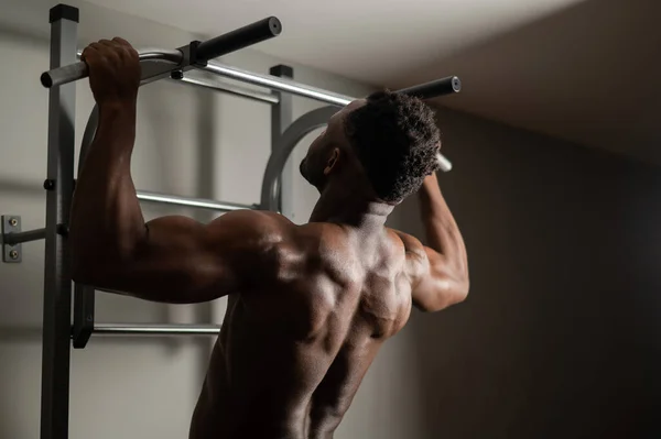Africain américain avec torse nu tire vers le haut sur barre horizontale dans la salle de gym. — Photo