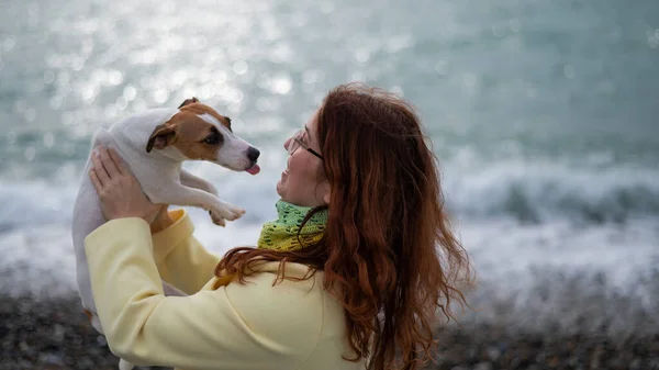 Mulher de cabelos vermelhos caucasiana segurando um cão jack russell terrier na costa. — Fotografia de Stock