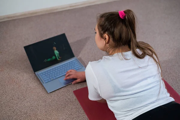 A chubby young woman is watching an online fitness lesson on a laptop. Distance sports training.