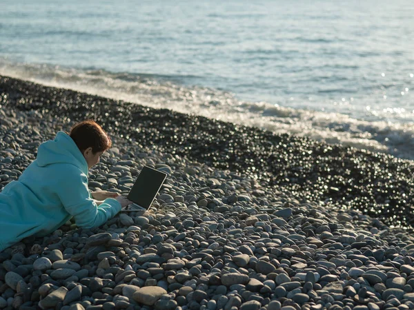 Beyaz bir kadın, çakıl taşı plajında yüzüstü yatıyor ve dizüstü bilgisayarında yazı yazıyor. Uzak çalışma serbest çalışanı.