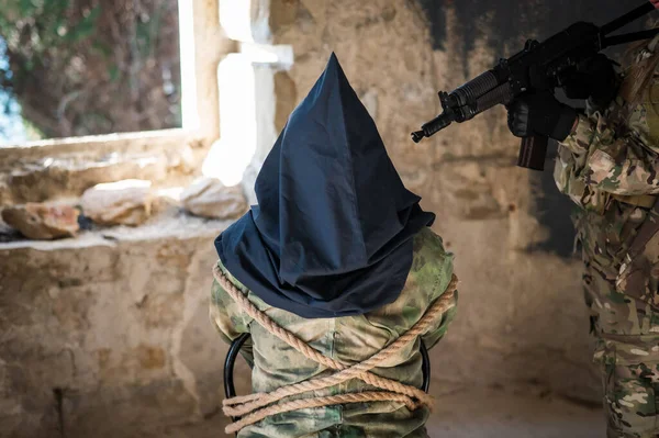 Een blanke vrouw in een legeruniform houdt een gijzelaar met een zak op haar hoofd bij het zien van een machinegeweer.. — Stockfoto