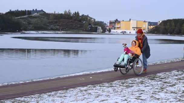 En kvinna i rullstol går med sin vän och en hund vid sjön på vintern. — Stockvideo
