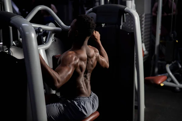 Bez košile africký Američan dělá zpět cvičení na stroji v tělocvičně. — Stock fotografie