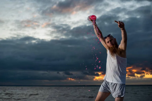 Um retrato bem-humorado de um homem brutal derramando refrigerante de uma garrafa na praia ao pôr-do-sol — Fotografia de Stock