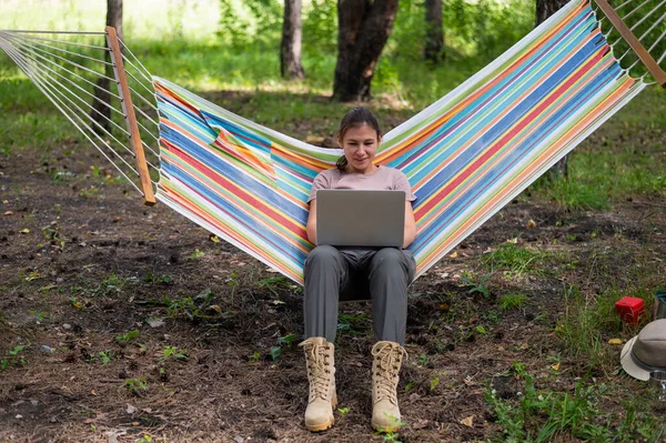 Mulher caucasiana trabalhando em laptop enquanto sentada em uma rede na floresta. Menina usa um computador sem fio em uma caminhada. — Fotografia de Stock