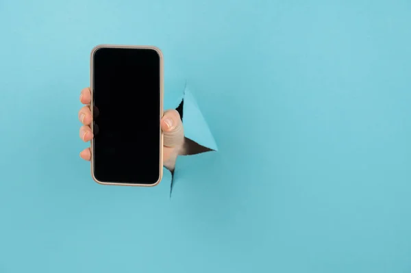 Una mano femenina que sobresale de un agujero de un fondo azul sostiene un teléfono inteligente con una pantalla negra. — Foto de Stock