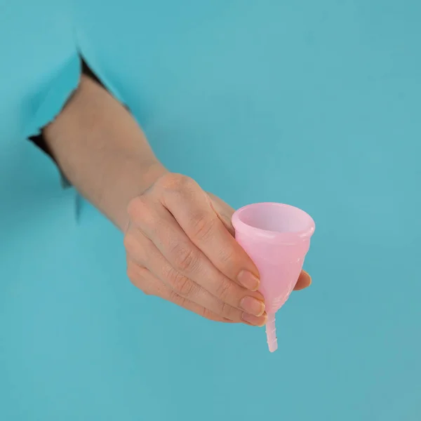 Una mano femminile che sporge da un buco su uno sfondo blu tiene una coppetta mestruale rosa. — Foto Stock